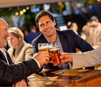 Three people seated at a wooden table in a bar, smiling and clinking their glasses of beer in a toast. The background shows other patrons and a dimly lit ambiance with string lights.