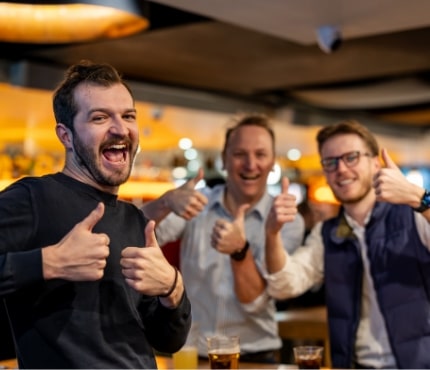 Three men are smiling and giving thumbs-up gestures in a well-lit bar or restaurant. They appear to be enjoying themselves, with drinks visible on the table in front of them. The background includes warm lighting and a busy atmosphere.