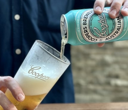 Person pouring a can of Coopers Brewery beer into a glass. The beer is amber-colored with a frothy head. The can and glass both have Coopers branding. The person is wearing a dark blue shirt, and the background is blurred.