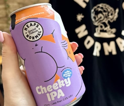 A hand holding a can of "Heaps Normal Cheeky IPA," a non-alcoholic beer. The can is purple with playful, abstract graphics and text. In the background, part of a shirt with a logo showing a dumbbell-lifting character and the words "Heaps Normal" is visible.
