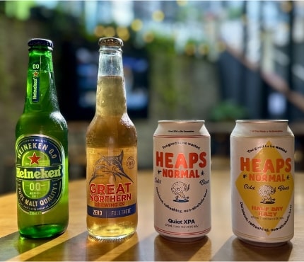 A close-up image of four beverages on a wooden surface. From left to right: a Heineken 0.0% non-alcoholic beer bottle, a Great Northern Zero Full Taste non-alcoholic beer bottle, a Heaps Normal Quiet XPA non-alcoholic beer can, and a Heaps Normal Half Day non-alcoholic beer can.