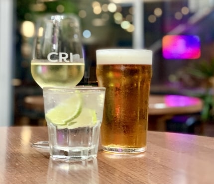 Three drinks are on a polished wooden table in a dimly lit setting: a glass of white wine with "CRI" on it, a pint of beer with a foamy head, and a cocktail glass with ice and lime wedges. The background is softly blurred with colorful lights.