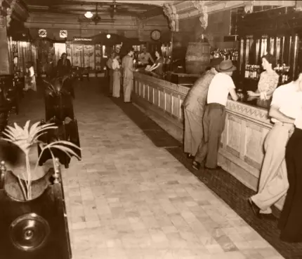 Sepia-toned image of a vintage bar with patrons and a bartender, featuring ornate decor, tile flooring, and high ceilings.