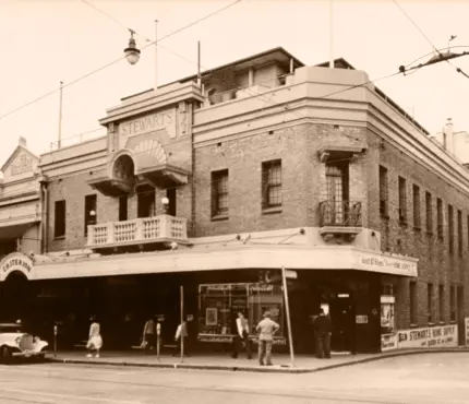 Vintage photograph of Stewart's theatre on a street corner, with people walking along the sidewalk and cars parked nearby.