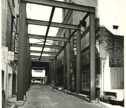 Black and white photograph of an urban alleyway framed with steel beams, featuring a lone person walking, brick buildings on both sides, and various signs and posters.