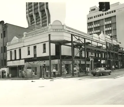 A partially constructed building on a street corner with pedestrians and cars in an urban setting, surrounded by older and modern buildings.