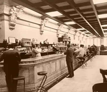 Vintage bar scene with patrons standing at a long counter, bartender on the other side, ornate columns and high ceiling in the background.