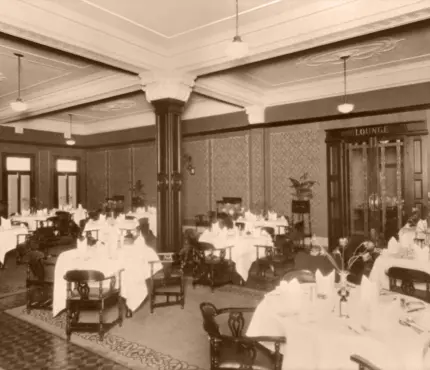 A vintage dining room with neatly arranged tables covered in white tablecloths, wooden chairs, and elaborate moulding on the ceiling.
