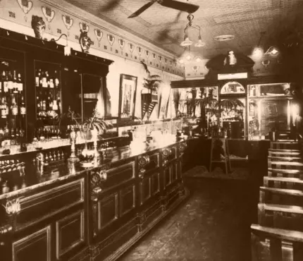 Vintage bar interior with wooden bar counter, bottles on shelves, ornate ceiling, and chairs along the side.