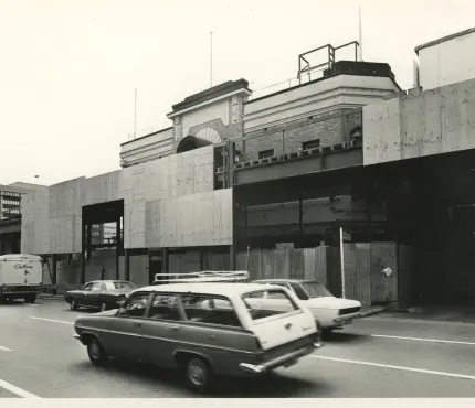 Black and white photo of cars driving past a partially demolishing building surrounded by wooden fencing.