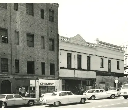 Black and white photograph of a street scene featuring older brick buildings, parked vintage cars, and a chemist store.