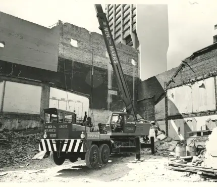 A crane labelled "MARRS" is positioned within a demolished building site, with a worker operating it amidst rubble and debris.