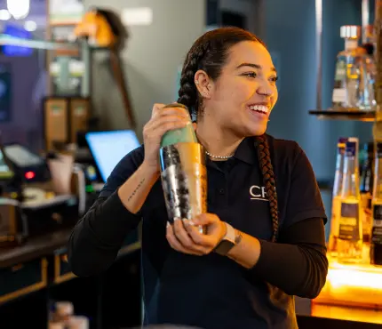 Bartender with braided hair shaking a cocktail shaker and smiling behind a bar.