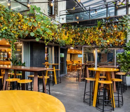 Outdoor seating area at a bar with tall tables and stools, greenery and string lights overhead.