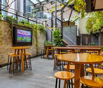 Outdoor seating area with wooden tables and stools, a large TV, and greenery hanging above and around the space.