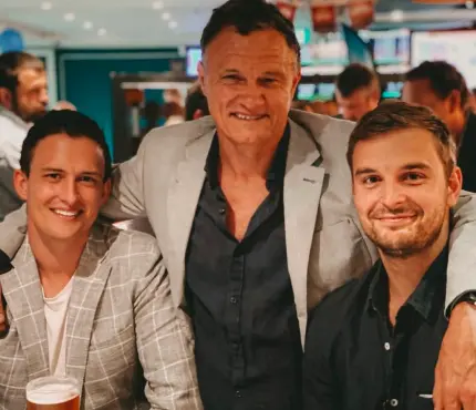 Three men smiling and posing together in a lively indoor setting, all dressed in smart casual attire.