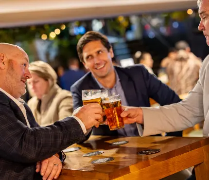 Three men in suits toasting with beer glasses at a wooden table in a bar.