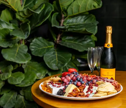 Cheese platter with assorted cheeses, fruits, nuts, and crackers, accompanied by a bottle of champagne and an empty flute glass on a wooden table with green foliage in the background.