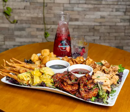 A platter of assorted appetisers including dumplings, skewers, spring rolls, and chicken wings, served with dipping sauces on a wooden table. A carafe of berry drink is in the background.