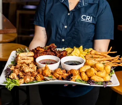 Person holding a large platter with various appetisers including wings, dumplings, and dipping sauces.