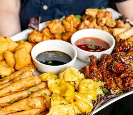 A platter with various Asian appetisers including fried dumplings, spring rolls, chicken wings, and dipping sauces.