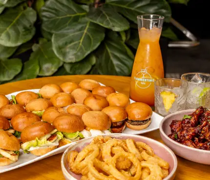 A platter of small burgers, a bowl of onion rings, a bowl of glazed chicken wings, and a tall pitcher of orange juice with two glasses on a wooden table.